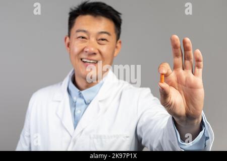 Lächelnder koreanischer Arzt mittleren Alters in weißem Mantel zeigt auf grauem Hintergrund isolierte Medikamentenkapsel Stockfoto