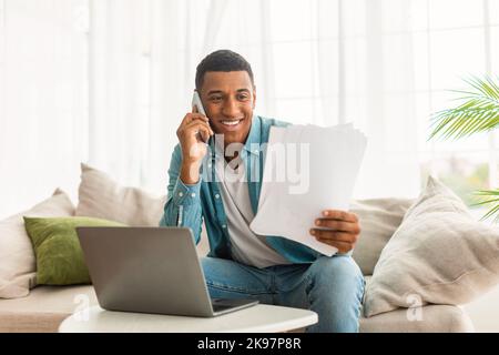 Fröhlicher junger schwarzer Mann in Jeans ruft per Telefon an, arbeitet mit Dokumenten und Laptop, liest Vertrag Stockfoto