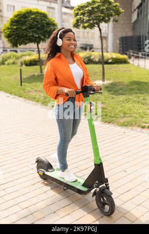 Glückliche tausendjährige afroamerikanische Dame in lässigen, kabellosen Kopfhörern, fährt auf einem Elektroroller Stockfoto