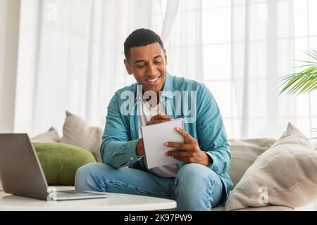 Lächelnder junger hübscher schwarzer Mann Student in Jeans macht Notizen, arbeitet mit Laptop Stockfoto