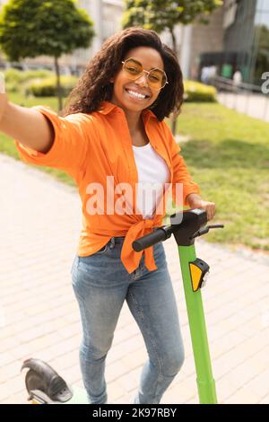 Happy Millennial african american Lady in Casual unter Selfie auf dem Smartphone auf Elektroroller in der Stadt Straße Stockfoto