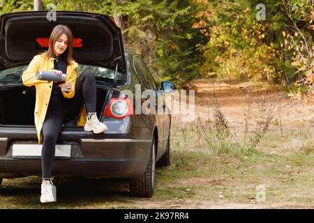 Die junge Frau sitzt im Kofferraum des Autos und gießt Tee aus einem Thermos im Herbstwald. Reisekonzept Stockfoto