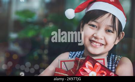 Das kleine Mädchen im Weihnachtshut lächelte hell, als sie die festliche Geschenkbox „Weihnachten“ und den Neujahrstag erhielt Stockfoto