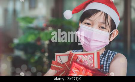 Das kleine Mädchen mit Gesichtsmaske und Weihnachtsmütze lächelte hell, als sie die festliche Geschenkbox „Weihnachten“ und den Neujahrstag erhielt Stockfoto