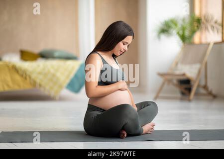 Junge Schöne Frau Erwartet Baby Auf Yoga-Matte Sitzen Und Umarmen Bauch Stockfoto