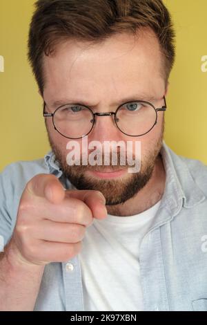 Der Mann mit Bart und Brille sieht streng aus und zeigt die Finger auf einen gelben Hintergrund. Stockfoto