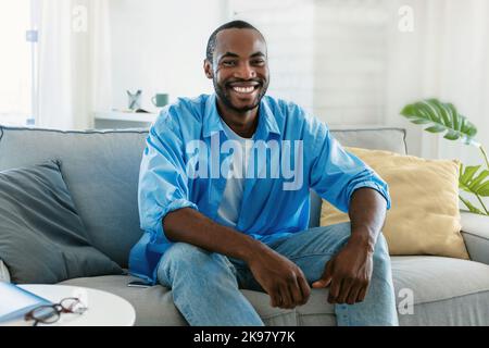 Porträt eines hübschen schwarzen Mannes, der lächelt und die Kamera anschaut, zu Hause auf dem Sofa sitzt und Zeit im Haus verbringt Stockfoto