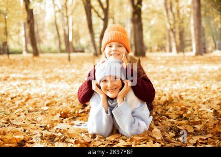 Lächelnde Frau und Mädchen in niedlichen Hüten, die auf dem Boden voller gelber, trockener Blätter aufeinander liegen. Glückliche Mutter und Tochter in gemütlichen Herbst-Outfits viel Spaß Stockfoto