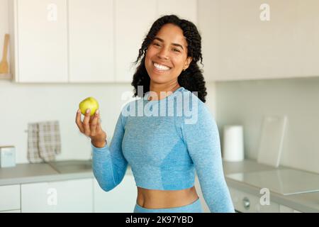 afroamerikanische sportliche Dame, die Apfel hält und die Kamera anlächelt, nach dem Training frisches Obst isst und in der Küche steht Stockfoto