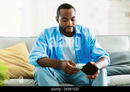 Der junge afroamerikanische Mann sitzt auf der Couch mit schwarzer Brieftasche in den Händen und zählt seine Ersparnisse, bevor er einkaufen geht Stockfoto