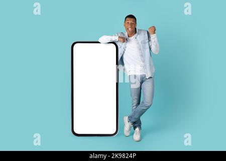 Glücklich aufgeregt tausendjährigen afroamerikanischen Mann in Jeans machen Sieg Geste mit der Hand in der Nähe von riesigen Smartphone Stockfoto