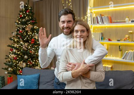 Porträt eines liebevollen, reiferen Familienpaares, eines Mannes und einer Frau, die sich umarmen und die Kamera betrachten, an Weihnachten in der Nähe des Weihnachtsbaums, um das neue Jahr zu Hause gemeinsam zu feiern. Stockfoto