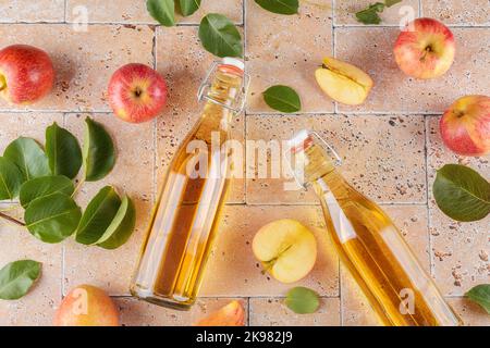 Apfelessig in zwei Glasflaschen und frische Äpfel mit Blättern auf beigefarbenem Beton-Hintergrund, Draufsicht, Nahaufnahme Stockfoto