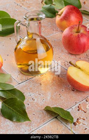 Apfelessig fermentierter Früchte in Glasflasche mit Äpfeln auf beigefarbenem Travertin-Tischhintergrund Stockfoto