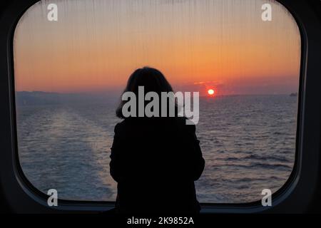 Denkende Frau, die während des frühen Morgensonnenaufgangs vom Fährenfenster aus auf das Meer blickt. Frau, die in einer Fähre mit Blick auf den Ozean und den Himmel unterwegs ist. Stockfoto