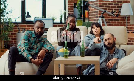Eine Gruppe von Freunden, die sich einen Actionfilm auf dem fernsehkanal ansehen und Bier aus Flaschen trinken. Menschen genießen Freizeitaktivitäten mit Getränken und Fernsehfilm oder Show. Stativaufnahme. Stockfoto