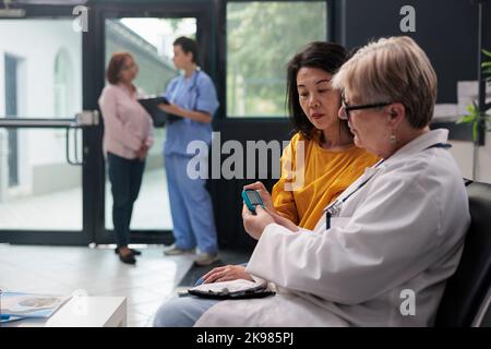 Leitender Arzt, der den asiatischen Patienten mit Glucometer konsultiert und den Insulin- und Glukosespiegel aus der Blutprobe misst. Medic und eine Frau, die bei der Untersuchung des Kontrollbesuches eine diabetische Kontrolle durchführen. Stockfoto