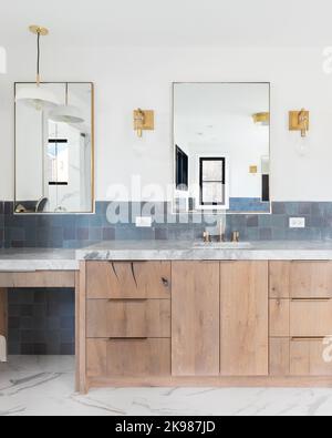 Das renovierte Badezimmer eines Luxushauses mit blauen Fliesen, einem Schminkschrank aus hellem Holz, goldfarbenen Lampen an der Wand und einer Arbeitsplatte aus grauem Stein. Stockfoto