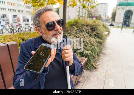 Eleganter, blinder, grauhaariger, reifer Mann mit Bart und dunkler Sonnenbrille, der einen Spazierstock hält, die Herbstsaison im Park genießt und über ein Freisprechtelefon mit jemandem spricht. Hochwertige Fotos Stockfoto