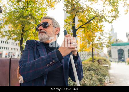 Blinder bärtiger grauhaariger reifer Mann mit dunkler Brille und einer Jacke, die einen Spazierstock hält und auf einer Bank im Park sitzt. Hochwertige Fotos Stockfoto