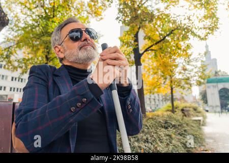 Eleganter, blinder, grauhaariger, reifer Mann mit Bart und dunkler Sonnenbrille, der einen Spazierstock hält und die Herbstsaison im Park genießt. Hochwertige Fotos Stockfoto