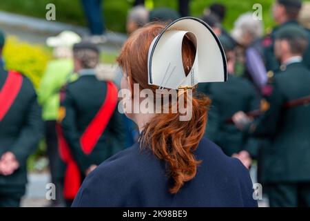 Eine junge Dame steht bei einer Militärparade mit einer Krankenschwester-Mütze und langen, leuchtend roten Haaren in einem Pferdeschwanz zurück. Es gibt Armeepersonal in Uniform. Stockfoto