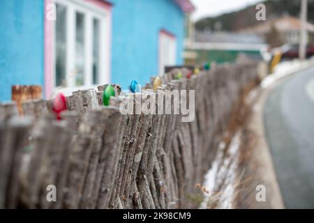 Ein rustikaler, ineinander greifender Zweigzaun aus Holz mit bunten Weihnachtslichtern auf der Oberseite. Eine Straße auf der einen Seite und ein blaues Haus dahinter. Stockfoto