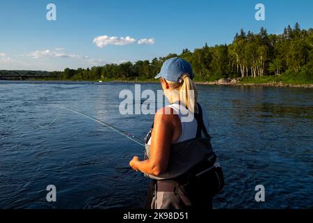 Eine Frau mittleren Alters steht in einem großen Lachsfluss und wirft eine Angelrute in ihre linke Hand. Die blonde, langhaarige Dame trägt ein Blau Stockfoto