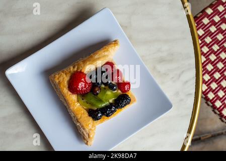 Ein viereckiges, mit Früchten gefülltes französisches Blätterteig auf einem weißen viereckigen Teller. Auf dem mit Sahnehühnchen gefüllten Dessert befinden sich Erdbeeren, Kiwi und Brombeeren Stockfoto