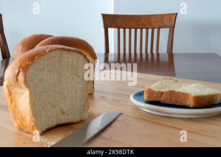 Ein einzelner Laib mit frischem, weißem, gekrustetem Brot auf einem Holzbrett auf einem Küchentisch. Das warme knusprige Brötchen hat die Butter über die knusprigen Brötchen geschmolzen. Stockfoto