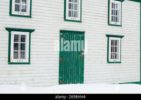 Die Außenwand eines alten weißen Holzgebäudes mit hellgrünen Zierleisten. Es gibt eine kleine Holztür und drei Glasfenster im Landhaus. Stockfoto