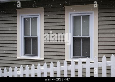 Weißer Pfostenzaun auf einer Diagonale im Vordergrund mit einem bräunlichen Haus im Hintergrund. Es gibt zwei weiße, doppelt aufgehängte Fenster mit beigefarbenen Zierleisten Stockfoto