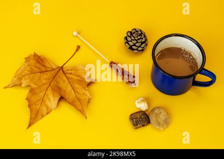 Eine Tasse heiße Schokolade mit abgefallenen Blättern, einer kleinen getrockneten Ananas, geröstetem Rohrzucker und Steinen auf hellgelbem Hintergrund Stockfoto