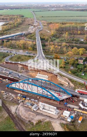 26. Oktober 2022, Sachsen, Großdeuben: Bauarbeiter bereiten sich auf eine Stahlbogenbrücke über die ehemaligen B95 und die zukünftigen A72 zwischen Leipzig und Chemnitz vor. Zwischen Oktober 28 und Oktober 31 soll die 79-Meter-Brücke am künftigen Anschlusspunkt Zwenkau oberhalb der neuen Autobahn ihren Platz finden. Mit Hilfe eines Schwerlastkran wird die rund 1.650 Tonnen schwere Konstruktion endlich auf sogenannten Aufbauten in ihre endgültige Position gebracht. Die Bundesstraße muss für dieses Wochenende komplett gesperrt sein. (Luftaufnahme mit Drohne) Foto: Jan Woitas/dpa Stockfoto