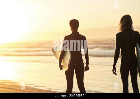 Die perfekte Welle wartet auf uns da draußen. Rückansicht eines jungen Paares, das am Strand surft. Stockfoto