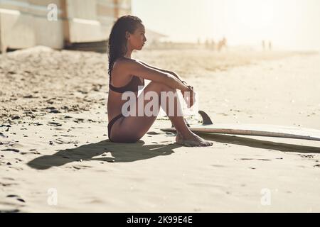 Ich bin gekommen, um all dem zu entkommen. Eine schöne junge Frau, die mit ihrem Surfbrett am Strand saß. Stockfoto
