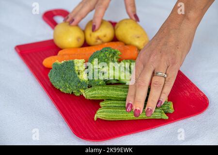 Frau bereitet Tisch mit frischen Salatzutaten zu. Zucchini, Kohl, Karotten und Kartoffeln auf einem roten Kunststofftablett Stockfoto