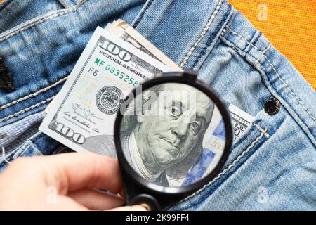 American Hundred Dollars ragen aus einem Loch in Jeans-Nahaufnahme, vergrößert mit einer Lupe zum Lesen Stockfoto