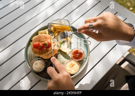 Nahaufnahme eines Burgers auf einem Teller und Hände eines unbekannten kaukasischen Mannes bereiten sich vor, an sonnigen Tagen im Freien auf dem Tisch zu essen. Im Restaurant halten Sie Gabel und Messer Stockfoto