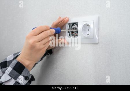 Nahaufnahme der Hände einer Elektrikerin, die einen Schraubendreher hält, der an einem elektrischen Stecker an einem elektrischen Wohnsystem arbeitet und eine weiße Steckdose installiert o Stockfoto