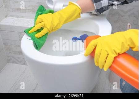 Nahaufnahme Hand mit Waschmittel Reinigung Toilettenschüssel im Badezimmer. Mann in gelben Gummihandschuhen, der Toilettensitz mit grünem Tuch reinigt. Badezimmer und Toilette Stockfoto