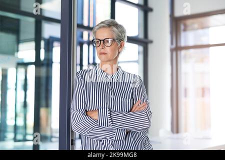 Ich muss hier einige ernsthafte Veränderungen vornehmen: Eine attraktive reife Geschäftsfrau, die mit gefalteten Armen in ihrem Büro posiert. Stockfoto