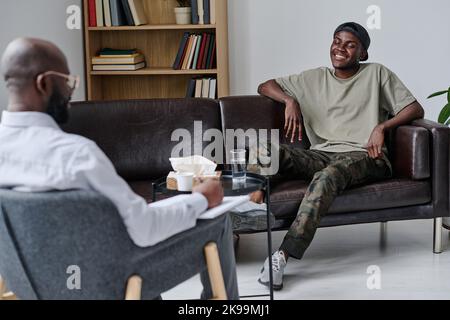Afrikanischer glücklicher Kerl, der auf dem Sofa sitzt und mit dem Psychologen im Büro spricht Stockfoto