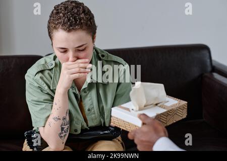 Mädchen mit Behinderung weint während einer Psychotherapie, während sie auf dem Sofa sitzt und eine Psychologin ihr Gewebe erreicht Stockfoto