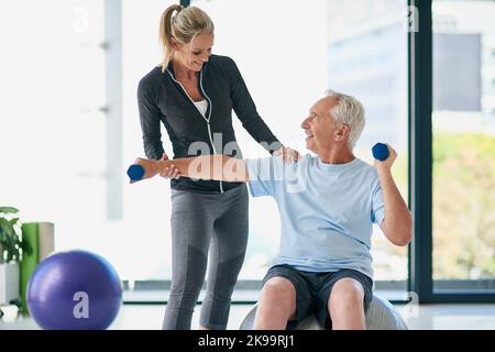 Ihre Muskeln werden stärker. Eine freundliche Physiotherapeutin hilft ihrer älteren Patientin mit Gewichten beim Training. Stockfoto