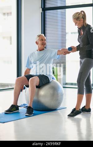 Lassen Sie uns versuchen, diese für ein paar Sekunden zu halten. Ganzkörperaufnahme eines glücklichen älteren Mannes, der mit Hilfe eines Physiotherapeuten mit Gewichten arbeitet. Stockfoto