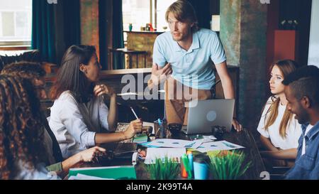 Der enthusiastische CEO spricht mit seinem Team von Fachleuten während eines Meetings im Loft-Stil, während die Mitarbeiter ihm zuhören und lächeln. Teamwork und Managementkonzept. Stockfoto