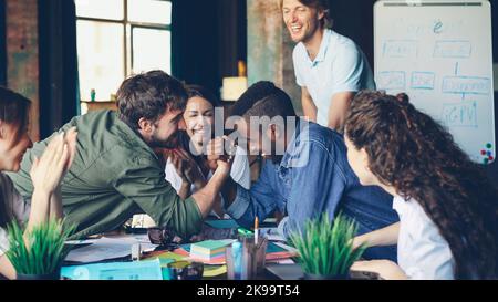 Fröhliche Kollegen, ein multiethnisches Team, üben während der Pause am Schreibtisch im Büro Armdrücken, Kollegen unterstützen sportliche Jungs Stockfoto