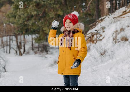 Glückliches Kind hält in den Händen Schneeball auf dem Hintergrund des verschneiten Waldes. Lächelnder Junge in warmer Kleidung, der Spaß im Freien hat. Konzept der weihnachtsferien Stockfoto