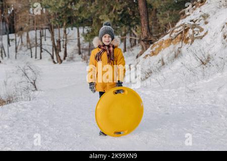 Happy Fun Kind hält einen Plastikschlitten auf dem Hintergrund des verschneiten Waldes. Junge, der Spaß an den weihnachtsferien hat. Konzept des Wintersports, aktive Freizeit Stockfoto